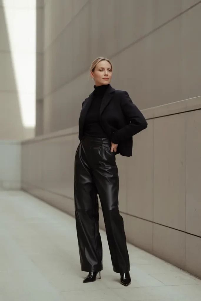 Full-length fashion portrait of a woman wearing a sophisticated all-black monochromatic outfit, featuring a fitted black blazer over a turtleneck and wide-leg black leather trousers, standing against a minimalist concrete architectural background