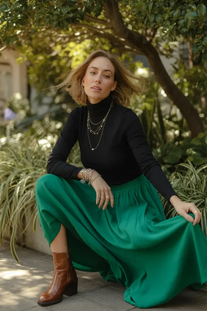 Full-length fashion portrait of a woman crouching in a sunlit garden, wearing a fitted black turtleneck and a flowing emerald green midi skirt, accessorized with layered gold necklaces and brown leather ankle boots.